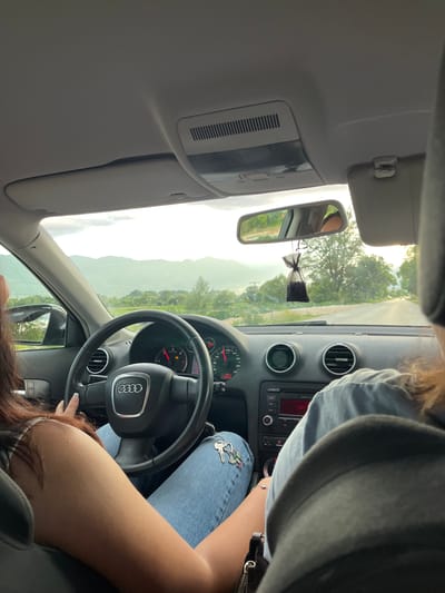 The image shows the interior of a car traveling on a road, likely near Dabnitsa, Bulgaria, given the mountainous landscape in the background. The perspective is from the back seat, looking forward. A woman with long, dark hair is driving, wearing a white and black striped top and blue jeans. Her left hand is on the steering wheel, which has the Audi logo. A set of keys with a charm is hanging from the gear shift. In the passenger seat, another person's shoulder and head, with long, reddish-brown hair, are visible. The dashboard features a radio and air vents. A small, dark air freshener is hanging from the rearview mirror. The road ahead is paved, with trees and vegetation on either side, and a hazy mountain range in the distance. The sky is overcast with some sunlight filtering through.