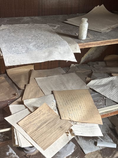 The image shows a drawer filled with old, dusty papers and a small bottle. The papers appear to be documents, some handwritten, with possible medical information in Ukrainian. The environment suggests abandonment and decay. The presence of the bottle and documents indicates a possibly medical or official setting. Pryp'yat', Ukraine is not evident in the image, although the Ukrainian language on the documents could suggest it.