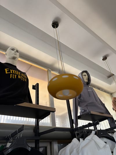 In Acıpayam, Turkey, this image captures an indoor retail environment, likely a clothing store, focusing on mannequins displayed on tiered shelving. To the left, a male mannequin is dressed in a black sweatshirt with yellow lettering that reads "Emirate FLY BETTER". To the right, another male mannequin is wearing a grey hooded sweatshirt. Suspended from the ceiling are two pendant lamps; one is a distinctive yellow, mushroom-shaped fixture with white polka dots, and the other is partially visible. The store features exposed white ceiling beams and metal shelving units. Below the displayed mannequins, racks of clothing, including several white shirts, are visible on hangers.