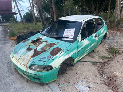 The image shows a dilapidated, light green and white, four-door hatchback car for sale, located outdoors. The car is heavily rusted, particularly on the hood, and missing wheels, exterior trim, and window glass. The driver's side door is open. A "CAR FOR SALE" sign, priced at 35,000 THB (Thai Baht), is displayed on the windshield. The car is situated on a gravelly or sandy surface with scattered debris and some vegetation. Background features include trees, a partial view of a building or structure, and the presence of grass. The scene appears to be in Baan Wang Ta Kien, Thailand.