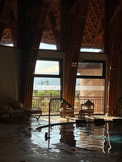 The image captures an inviting indoor space, likely within a resort or lodge in Mu Cang Chai, Vietnam. The scene is dominated by natural materials, with walls and ceilings constructed from intricately arranged bamboo or wooden slats, forming various geometric patterns. This rustic yet sophisticated design element creates a warm and earthy ambiance.
Large, dark-framed windows or glass doors open up to a bright outdoor view featuring lush green foliage and a clear, blue sky, indicative of a serene natural landscape. Through one of the windows, a blind with a distinctive white vertical strip adorned with small, repeating decorative motifs is visible.
In the foreground and extending to the right, a significant body of water, presumably an indoor or semi-indoor swimming pool, reflects the surrounding environment. The rippling surface beautifully distorts the reflections of the bamboo structures and the outdoor scenery into abstract patterns of light and shadow. A metal handrail or piece of equipment is positioned near or within the water.
Wooden furniture, including chairs with light-colored cushions, is arranged near the windows, suggesting comfortable seating areas for relaxation. No people are present, contributing to the tranquil and undisturbed atmosphere. The bright sunlight streaming through the windows and reflecting off the water indicates a daytime setting, likely mid-morning or afternoon, under clear and sunny weather conditions. The overall impression is one of peace, relaxation, and harmony with nature, consistent with a retreat in a scenic location like Mu Cang Chai. No discernible text is visible within the image.