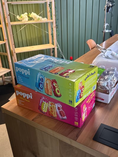 Here is a detailed description of the image:
**General Description:**
The image shows a close-up of a wooden table surface on which two boxes of "Poppi" sparkling prebiotic soda are placed. The boxes are situated next to a pack of glass bottles, seemingly containing a similar beverage. The background includes a wall with horizontal panels and a wooden structure with some floral decorations.
**Detailed Analysis:**
* **Foreground:**
* Two boxes of "Poppi" soda are the primary focus. One box, predominantly blue and green, is labeled "Everyday Icons." The other box, pink and orange, is labeled "Juicy Faves." Both boxes indicate that they contain 18 cans of soda.
* Behind the Poppi boxes, there is a clear plastic package containing glass bottles.
* A small black rectangle, possibly a coaster or a decorative element, is visible on the upper right corner of the table.
* **Background:**
* The wall in the background is made of wooden panels arranged horizontally. The panels appear to be painted in a muted green or grayish-green color.
* A wooden structure, possibly a trellis or part of a decorative installation, is partially visible. It has a grid-like design.
* White floral decorations are attached to the wooden structure.
* **Environment and Lighting:**
* The scene appears to be indoors, possibly in a studio or a staged area.
* The lighting seems to be soft and diffused, with no strong shadows.
* **Additional details:**
* There are some lighting equipment in the top left corner.
* It is evident that the scene has been rotated, as the horizontal wall panels appear to be vertical.
**Location:**
Given the presence of Poppi soda, a product commonly available in the United States, the scene likely takes place in the USA. Without more details, the exact location cannot be determined solely from the image.