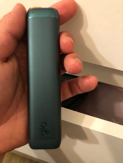 A person's hand is holding a teal-colored electronic smoking device with the logo "glo" embossed on its side. The device has a bright orange tip. The background appears to be a white box or surface, with a portion of the device partially visible within it. The image is a close-up shot, focusing on the device and the hand. No specific location in Leszno, Poland can be determined from the image.
