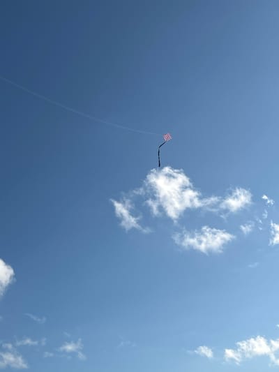 In a clear, blue sky, a kite flies high, its diamond-shaped body adorned with a pink and white checkered pattern. A long, dark tail trails behind it. To the right of the kite, a fluffy white cloud drifts by, its edges softened by the sunlight. Further down and to the left, another smaller cloud can be seen. In the far distance, a faint white line, likely a contrail from an airplane, stretches across the sky. The scene is set against a vast expanse of blue, suggesting an open outdoor space, possibly a park or field in the city of A R A, Indonesia, where a kite-flying activity is taking place.