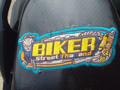 The image is a close-up, eye-level, medium shot of an embroidered patch on a black, slightly textured surface, possibly a motorcycle part. The patch is irregularly shaped and features the word "BIKER" prominently in large, bold, yellow letters outlined in dark blue and black. Below "BIKER," smaller white text reads "Street Tha land," also outlined in dark blue and black. The patch is surrounded by a bright turquoise border. Various illustrative elements are incorporated into the design, including what appears to be a stylized motorcycle engine or mechanical components in shades of gray, white, yellow, and orange. The background surface is mostly black with some visible texture and a few small imperfections or specks. The lighting is even, highlighting the detail of the embroidery. The location in the city of Megamendung, Indonesia, is not visually discernible from the provided image.