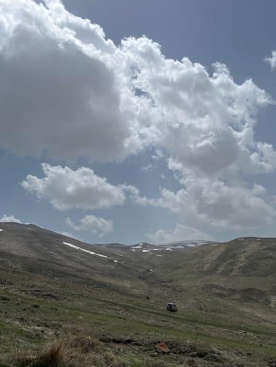 The image depicts a mountain landscape in Hartagyugh, Armenia, under a partly cloudy sky. A mountain range stretches across the right side of the frame, showing patches of snow near the ridgeline. Vegetation covers the lower slopes, with varying shades of green and brown. A single vehicle is parked on the slope of the mountain. The sky occupies the left portion of the image, filled with large, white cumulus clouds against a backdrop of light blue sky. The image is rotated so that the mountain and vehicle are positioned on the side.