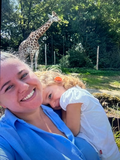 A selfie-style image shows a woman and a young child, possibly at a zoo in Braintree, United Kingdom. The woman, wearing a blue shirt, is smiling. The child is leaning on her, wearing a white shirt and appears to be looking into the camera. A giraffe is visible in the background, to the left of the frame. Trees and vegetation are also visible in the background. A fence or structure is also visible behind the giraffe. The scene appears to be outdoors in daylight.