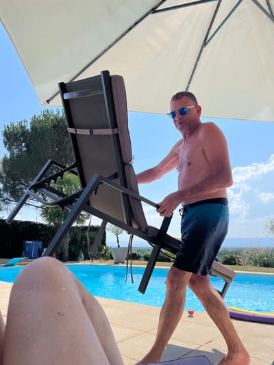 In Saint-Galmier, France, a shirtless man with sunglasses stands near a swimming pool, holding a reclining chair under a large umbrella. He is wearing blue and black swimming trunks. The swimming pool is filled with bright blue water, and the surrounding area is paved with light-colored tiles. Lush green trees and vegetation are visible in the background, along with a distant hilly landscape under a clear blue sky with scattered white clouds. The scene appears to be a sunny day at a private residence or resort.