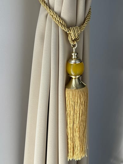 The image shows a close-up view of a golden curtain tie-back with a tassel. The tie-back is made of a thick, braided gold rope that is looped around and knotted to hold a beige curtain. Hanging from the knot is a decorative element consisting of a gold-colored finial, a translucent yellow orb, and a long, golden tassel. The curtain itself appears to be made of a textured fabric and is drawn to reveal a grey wall in the background. The lighting suggests an indoor environment. There are no people or animals present in the image. The image does not contain any information that would allow for a specific location in Tanjungmeru, Indonesia.