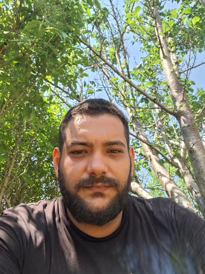 A close-up, outdoor, waist-up selfie features a man with a dark beard and short dark hair looking directly at the camera. He is wearing a black t-shirt. He is positioned in front of lush green foliage and tree trunks, with a clear blue sky visible through the branches. The scene appears to be a park or a garden area in Dolna Malina, Bulgaria.