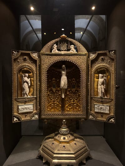 The image shows a reliquary, a decorative container for religious relics, displayed in a museum setting. The reliquary is made of gold and intricate metalwork, with enamel inlays and carved ivory or stone figures. At the center, a figure of Christ on the cross is depicted. Flanking the central panel are two hinged doors, each containing a carved figure of an angel and a smaller scene at the bottom. The reliquary stands on an ornate base. The background is dark, with reflections visible in a glass display case, suggesting an interior exhibition space. The style of the reliquary suggests it is a historical artifact, likely religious in nature, and possibly from the medieval or Renaissance period. The reliquary is part of the collection of the Museo Catedralicio (Cathedral Museum) of Santiago de Compostela, Spain, which houses treasures from the Cathedral of Santiago de Compostela, a major pilgrimage site.