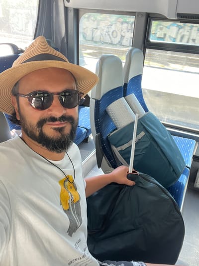 A man in a straw hat and sunglasses is taking a selfie on a bus. He is wearing a white t-shirt with a graphic print. Next to him, on the bus seat, are two rolled-up sleeping mats inside a teal bag and a large dark bag. The bus windows show a view of graffiti and train tracks, suggesting the bus is traveling through an urban area. This scene is consistent with a location in Montcada i Reixac, Spain, known for its industrial areas and graffiti along railway lines.