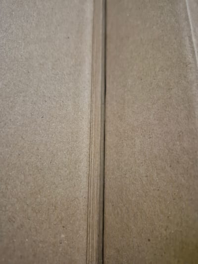 The image shows a close-up of a brown cardboard surface. The texture of the cardboard is visible, with small fibers and specks. A vertical crease or seam runs down the center of the image, dividing the surface into two sections. The lighting appears to be diffused, creating soft shadows and highlighting the texture of the material. There are no people, objects, or actions depicted in the image, and no specific location in Pabradė, Lithuania, can be identified.