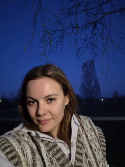 A selfie shows a woman with light skin, brown hair, and green eyes, wearing a brown and white sweater over a white shirt. She is smiling slightly. Behind her is a dark blue sky with trees silhouetted against it, with branches displaying new spring leaves. Two small lights are visible in the distance. It is not possible to determine the exact location of the photo, even given the city is Узяр, Russia.