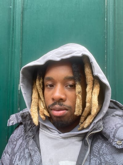 A man with dreadlocks, partially blonde, wearing a gray hoodie and a gray and black jacket with a snakeskin pattern, is looking directly at the camera. He has brown eyes and a slight beard. The background is a green, textured surface. The picture was most likely taken indoors. I cannot verify the location in the city of Grenoble, France.