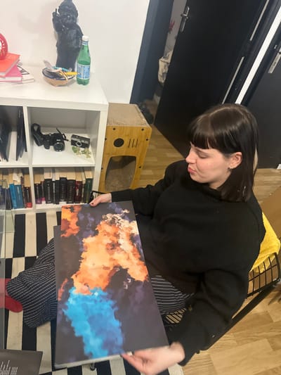 A person with dark hair and a lighter streak looks down at a rectangular print featuring an abstract cloudscape in shades of orange, blue, and black. They wear a black sweater and striped pants. To the left, a white shelf holds a cat condo, books, a camera, and various objects. The room has a black and white striped rug and a wooden floor.