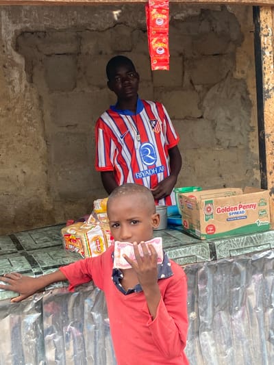 A medium shot shows two young boys in front of a market stall in Rigachikun, Nigeria. The boy in the foreground, wearing a red shirt with a dark blue collar, holds a wad of Nigerian Naira bills to his mouth. He has short, dark hair and is looking directly at the camera. Behind him, a slightly older boy, wearing a red, white, and blue striped athletic shirt with "RIYADH AIR" printed on it, stands in the stall. The stall is constructed of rough, light-colored bricks and has a wooden frame at the top. Red packets of tea are hanging from the top of the stall. Several loaves of bread wrapped in plastic are visible on the counter, along with a box labeled "Golden Penny SPREAD." The counter surface appears to be tiled in a green and grey pattern. The background is a rough, unpainted brick wall, and the foreground features a corrugated metal surface.