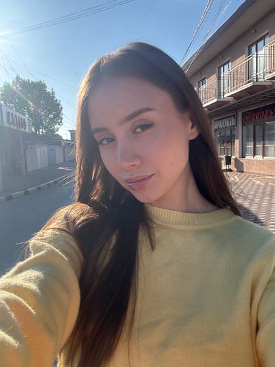 A woman is standing outside in a location near Береговой сельский совет, Ukraine. She is wearing a light yellow sweater. The sky is blue and sunny. In the background is a street with buildings and trees. There are power lines visible. The woman is holding the camera at an angle, taking a selfie.