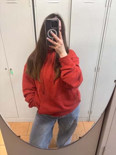 A person is taking a mirror selfie in what appears to be a locker room. The person is wearing a red hooded sweatshirt and light blue jeans. They are holding a smartphone with a magnetic ring on the back. In the background, there are white lockers and a tiled floor. The environment suggests a public facility like a gym, school, or community center.