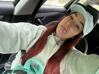 A young woman with red hair and freckles is taking a selfie inside a car. She is wearing a light blue hooded sweatshirt and a light blue visor. She is sticking her tongue out. A black seat and seatbelt are visible, as is a light blue backpack. The location in the city of Novosellë, Kosovo is not discernible.