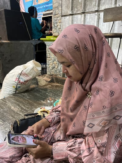 An Indonesian woman wearing a pink patterned hijab and matching dress is sitting at a wooden table, looking at her phone. The phone screen displays a picture of a woman's face wearing a hijab. A black handbag sits on the table next to her. In the background, a man in a teal shirt is working on some equipment near a large speaker. A white sack with text printed on it, possibly fertilizer, is leaning against a stone wall. The setting appears to be outdoors or in a covered open-air space, with corrugated metal and stone walls visible. The overall scene suggests a casual gathering or event. The image is taken in the city of Wedarijaksa, Indonesia.