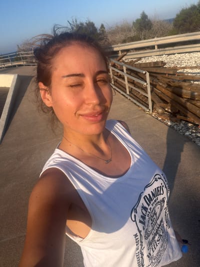 A woman with dark hair tied up in a messy bun, wearing a white tank top with a Jack Daniel's logo, is captured in a selfie on a paved walkway. She is winking with her eyes closed, and a subtle smile plays on her lips. A silver necklace with a pendant spelling "mama" adorns her neck. The background reveals a guardrail and a pile of wooden planks, suggesting a construction or renovation site. Beyond that, trees and the vast expanse of the sky are visible. The setting sun casts long shadows and illuminates the scene with a warm, golden light, indicating it's either sunrise or sunset. The environment appears to be outdoors, possibly near a coastline, given the clear sky and natural elements.