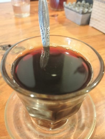 A clear glass of dark coffee with a silver spoon is placed on a wooden table. The coffee has a foamy surface with small bubbles. In the background, out of focus, is a glass of water and a small planter with decorative stones. The image is taken from a close-up, overhead angle, focusing on the coffee and spoon.
