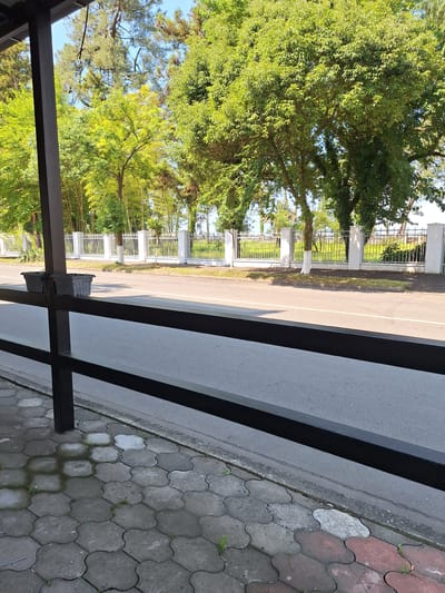 The image shows a street scene in Kobuleti, Georgia. In the foreground, a dark metal railing runs horizontally across the frame, separating a tiled walkway from a paved road. The walkway is made of interlocking hexagonal and clover-shaped gray and red concrete pavers. Two dark gray rectangular planters, one slightly further back than the other, sit on a dark vertical post to the left, partially obscured by the railing.

Beyond the road, a white metal fence with vertical bars and decorative white pillars marks the boundary of a property. Behind the fence, lush green trees, some with vibrant yellow-green foliage, are prominent. Some of these trees appear to be palms and bamboo. The sky above is clear and blue. The scene suggests a sunny day in a well-maintained, possibly residential or public area.
