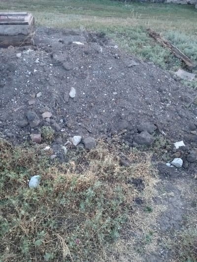 A pile of dark, damp soil and rubble sits in the foreground, with some dry, green grass and weeds growing around its base. To the left, a concrete structure with a metal handle is partially visible, embedded in the ground. A piece of wood lies on the ground to the right of the soil pile. The background consists of a grassy field.