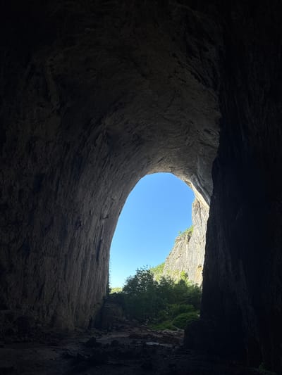 The image captures the view from inside the Prohodna Cave, also known as the "Eyes of God," in Karlukovo, Bulgaria. The perspective is from the dark interior of the cave, looking out towards a bright, open sky. The cave walls are massive and textured, with the opening forming a distinct, large archway.
Dominating the foreground is the rough, textured rock of the cave's interior, rendered in dark, shadowed tones. The scale of the rock formations is evident, showcasing their geological ruggedness. As the eye moves towards the center and right side of the frame, the darkness gives way to the immense arch of the cave's entrance.
Through this arch, a vibrant blue sky is visible. Beyond the immediate entrance, a lush green landscape unfolds, characterized by dense trees and foliage. The sunlight illuminates the surrounding cliffs, highlighting their layered rock structure. The contrast between the dark, enclosed space of the cave and the bright, open expanse outside is striking.
In the lower right section of the cave floor, near the transition to the outside, a few silhouetted figures are visible. They appear to be small in comparison to the cave's scale, suggesting they are visitors or individuals exploring the area. The ground inside the cave is uneven, composed of rocks, dirt, and debris, indicative of a natural, untamed environment. The overall scene evokes a sense of awe and the grandeur of nature.