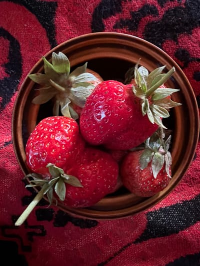 A close-up, overhead shot captures a brown, shallow bowl filled with several bright red strawberries, their green leafy tops visible. The bowl rests on a textured fabric with a bold red and black striped pattern. The image is well-lit, highlighting the glossy surface of the strawberries and the details of their seeds. The setting appears to be indoors, likely a kitchen or dining area, but specific location details in Тимоново, Russia, are not discernible from the visual information provided.