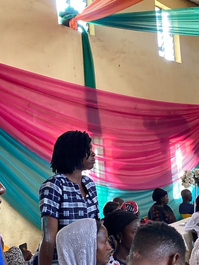 The image depicts an indoor gathering, likely a ceremony or event, set within what appears to be a community hall or place of worship in Turu, Nigeria. The background is dominated by draped fabric in vibrant shades of pink, teal, and orange, creating a festive and colorful ambiance. Sunlight streams through a window in the upper right corner, casting a bright glow and illuminating the space.
In the foreground, a woman with dark, braided hair is the central subject, facing to the right in profile. She is wearing a black and white checkered dress. To her left, a portion of another individual's face is visible. The middle ground features a gathering of people, mostly women, seated or standing, many wearing headscarves. A table is set in the background with a floral arrangement. The overall scene suggests a communal event, possibly a wedding, funeral, or religious service, characterized by its colorful decorations and the presence of a congregation.