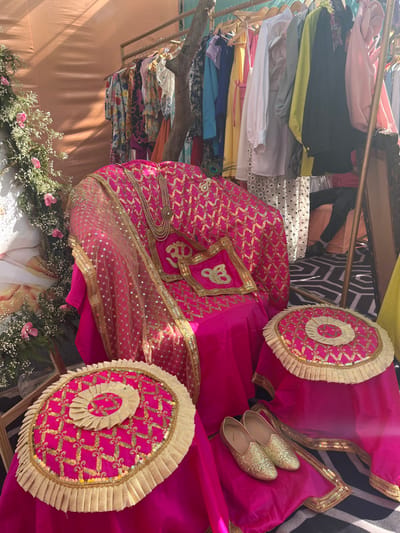 The image shows a close-up of a decorative wedding setup in a vibrant, outdoor setting. The scene is located in Sector 17, Chandigarh, India, at coordinates 30.764 latitude and 76.797 longitude. The centerpiece is a pink, sequined chair draped with a matching pink shawl adorned with gold embroidery. Two pink, sequin-covered cushions with gold embroidered accents are placed on the chair, and a small, ornate necklace rests on the seat.  Two additional pink cushions with gold embroidery and ruffles are situated on the floor. Two pairs of golden shoes are placed on the pink fabric to the right of the chair. The chair and cushions are arranged on a black and white patterned floor. A rack filled with colorful clothing is visible in the background.  The surrounding environment includes lush green foliage and a peach-colored wall. The image captures the elaborate decorative details often used in Indian weddings.