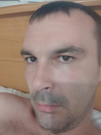 The image is a close-up, selfie-style photograph of a man's face and upper body, taken indoors. The man has short, dark hair, with a receding hairline. His skin is light-toned with some visible pores and a faint mustache and stubble. He has dark eyes and eyebrows. Part of his shoulder and chest are visible, showing light skin and some hair. The background is a light brown wooden surface, possibly a headboard or furniture. The lighting appears to be natural and soft, creating subtle shadows on his face. The image quality is moderate, with some graininess. There is no indication of the specific location within Kobeliaky, Ukraine.