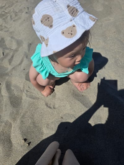 The image shows a baby sitting on a sandy beach, wearing a white sun hat with a teddy bear pattern and a turquoise swimsuit. The baby is viewed from above, with their legs tucked underneath them. The sand appears wet and reflects the sunlight, indicating it's a sunny day. In the background, there is a shadow cast on the sand, suggesting the presence of an adult or an object casting the shadow. The overall scene conveys a sense of a peaceful and relaxed moment on a beach, likely during the daytime. No visible text is present in the image.