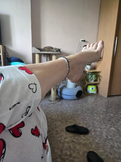 A person's left foot, adorned with a beaded anklet, is raised in the foreground, resting on a white garment patterned with red hearts. The foot is clean and appears to be of a pale complexion. The background is a domestic interior, with a cat sleeping on a cat tree, a shelving unit with plants and decorative items, and a closet door. Two black slippers are visible on the floor. The overall environment suggests a relaxed home setting. There is no specific indication of the city of Kimry, Russia.