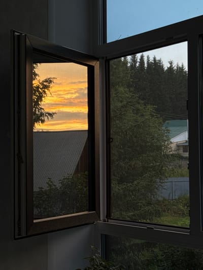 This image depicts an open window looking out onto a scene in Burdyata, Russia. The upper part of the window frames a view of dark green trees and some structures partially covered by a pale green tarp. The sky is a light blue. The lower part of the window is angled and reflects a vibrant sunset with streaks of orange and yellow clouds. Silhouetted against the sunset is the dark shape of a tree. To the right of the reflection, a grey, corrugated metal building is visible. The setting appears to be residential, given the presence of trees and buildings. The time of day is clearly sunset, indicated by the colors in the sky. The mood is tranquil and reflective, with the beauty of the sunset contrasting with the more mundane view of the surroundings. There is no visible text in the image.