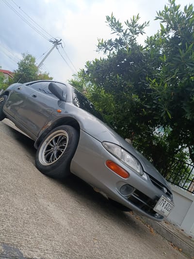 A grey, two-door coupe is parked on a concrete road. The car is viewed from a low angle, tilted upwards and to the right. The sky above is overcast, suggesting a cloudy or potentially rainy day. Lush green trees with dense foliage line the background, casting shadows. A power line pole with multiple wires is visible to the left of the car, extending into the sky.
The car appears to be a Honda Beat, a small, lightweight sports car. It has aftermarket alloy wheels with a black and silver finish. The front of the car shows a license plate with Thai characters and numbers, reading "กจ 6329" and some smaller text below. The car has a small orange turn signal light on the front fender. There are some signs of wear and tear on the car, particularly around the wheel arch, with visible rust.
The setting appears to be a residential area in Ban Patan, Thailand, given the presence of the car, the road, the greenery, and the style of housing hinted at by a white wall and fence in the background. The overall mood is ordinary, with no specific event or activity taking place; the car is simply parked.