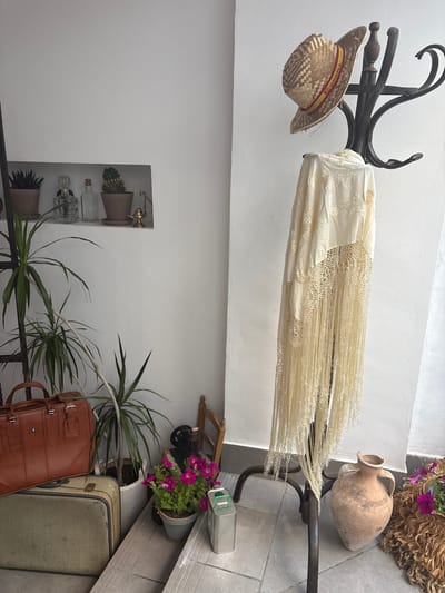The image shows an interior setting in Córdoba, Spain, with a rustic and slightly eclectic decor. A wrought iron coat rack holds a straw hat with the Spanish flag band, and a cream-colored shawl. A clay jug rests on the rack. In the background are plants, decorative bottles on a recessed shelf, a sewing machine, and stacked luggage. The environment includes a mix of white walls and tiled flooring, typical of Andalusian design. There are no people in the frame, only inanimate objects.