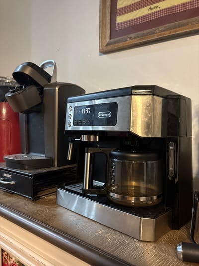 A black and silver Delonghi coffee maker is positioned on a patterned surface. The digital display reads "11:37". The coffee maker appears slightly dusty. A black Keurig machine is situated below and partially obscured. A framed item hangs on the wall to the left, displaying a patterned fabric with visible stitching and the words "Amen". The angle of the photo gives the impression that the objects are tilted.