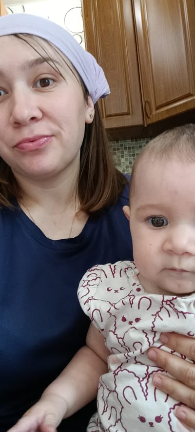 In a domestic indoor setting, a woman wearing a navy blue t-shirt and a light purple headscarf holds a baby. The baby is wearing a cream-colored onesie with a repeated pattern of stylized purple rabbits. The woman's face is partially visible in the upper left quadrant of the image, looking towards the camera with a slight smile. The baby's face is in the right half of the image, looking directly at the camera with a neutral expression. The background shows kitchen cabinets made of wood and a tiled backsplash. The overall scene suggests a casual, everyday moment within a home. There is no information in the image to indicate the specific location in the city of Хохлы, Russia.