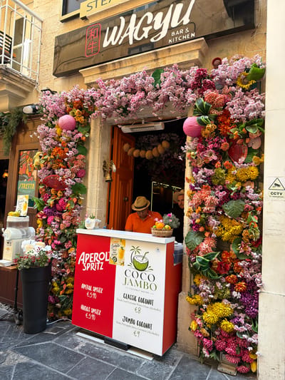 The image depicts an outdoor scene in Valletta, Malta, characterized by a vibrant floral arrangement adorning the entrance of a establishment. The flowers are a riot of colors, including pinks, oranges, yellows, and purples, creating a festive and inviting atmosphere.
A person wearing an orange shirt and a straw hat is visible behind a counter, serving drinks. The counter itself displays a menu with various beverage options, including "COCO JAMBO - COCONUT WATER," "CLASSIC COCONUT (€8)," and "JUMBO COCONUT (€9)." Below this, there is a section for "APEROL SPRITZ," with listings for "APEROL SPRITZ (€5.90)" and "HUGOS SPRITZ (€5.90)."
To the left of the entrance, a dark sign with white lettering reads "STEAK & BAR & KITCHEN," with what appears to be a red symbol in a black square. Further to the left, a window with a metal grate is visible.
The overall impression is one of a lively and colorful establishment, likely a restaurant or bar, with a focus on refreshing drinks and a welcoming ambiance. The bright floral decor suggests a sunny day, and the outdoor setting implies a pleasant time of day for people to be out and about. The scene evokes a sense of enjoyment and relaxation.