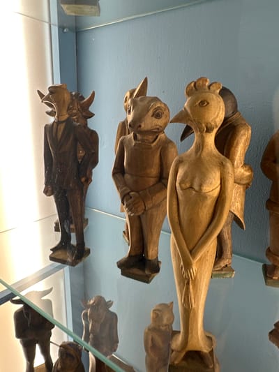 The image shows a collection of carved wooden figures displayed on glass shelves within a cabinet, likely a display case. The background is a light blue wall. The figures appear to be anthropomorphic animals or mythical creatures, each with unique characteristics.

In the foreground, a wooden figure with a bird-like head and a female torso stands with its hands clasped in front. To its left, a figure with a frog-like head and human-like body is also visible. Further to the left, a figure dressed in a suit with animalistic head and horns is present. Several other figures, partially obscured or in the background, also suggest animalistic or humanoid forms. Some of these figures are dark brown, while others are a lighter wood color.

The lighting suggests an indoor setting, possibly a museum or a private collection. The overall impression is of a display of handcrafted art objects. The specific location in Salzburg, Austria, cannot be determined from the image alone, as there are no identifying landmarks or contextual clues.
