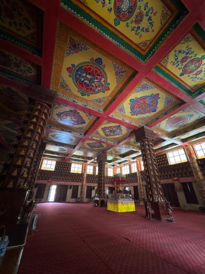 This image depicts the interior of a brightly colored, ornately decorated building, possibly a temple or cultural center. The ceiling is a dominant feature, divided into square panels with intricate yellow, red, green, and blue patterns, including floral motifs and circular designs. Large wooden columns, adorned with numerous small golden Buddha statues, support the ceiling and line the spacious room. The floor is covered with a rich red carpet. In the background, windows with natural light filtering through line the walls, and doorways are visible. A yellow altar-like structure is present in the mid-ground. The overall impression is one of grandeur and cultural significance. This image is not located in Việt Trì, Vietnam, as there is no visual evidence to support this claim.