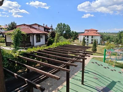 The image depicts an outdoor scene in Starosel, Bulgaria, under a blue sky with scattered white clouds. In the foreground is a wooden pergola, casting shadows on a green play area with black mounds and ropes. To the right of the play area, a playground structure is visible. In the midground, there are landscaped grounds with trees and bushes, and two buildings with red-tiled roofs are apparent. The building on the left is white with a dark-trimmed porch, while the building on the right is a multi-story structure. In the background, a tree-lined landscape with rolling hills is present. There are no people visible in the image.