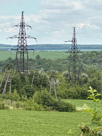 The image shows a rural landscape with two large electricity pylons dominating the foreground. The pylons are dark brown, lattice structures with multiple crossbars supporting thick power lines. They are positioned at varying distances from the viewer, with the left pylon appearing larger and closer than the one on the right.
In the mid-ground, a dense forest of green trees occupies the rolling hills. Interspersed within the trees are smaller, older wooden power poles, some of which appear weathered and partially obscured by foliage.
The foreground and background are characterized by agricultural fields. The immediate foreground features a lush, bright green field, likely planted with crops. Further in the background, there are more fields, some appearing a lighter shade of green, suggesting different types of vegetation or growth stages. The sky above is a pale blue with scattered, puffy white clouds, indicative of a daytime scene.
The scene is devoid of any people or animals. The overall impression is one of a functional, rural power infrastructure set against a backdrop of natural greenery and cultivated land. The specific location is the village of Lysohirka, Ukraine.