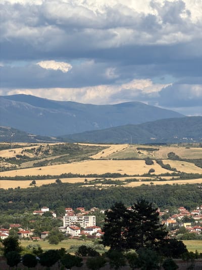 This is a landscape photograph taken in Kocherinovo, Bulgaria. The scene is divided into several horizontal planes. The uppermost plane is dominated by a dramatic sky filled with a mix of dark, heavy clouds and lighter, softer clouds. Sunlight breaks through in some areas, creating a sense of depth and contrast.
Below the sky, a series of rolling hills stretch into the distance. The farthest hills appear as faint, blue silhouettes, indicative of their distance. Closer hills are covered in lush green vegetation and trees.
In the mid-ground, vast fields of golden crops, likely wheat or barley, dominate the landscape, their color suggesting they are ready for harvest or have recently been harvested. These fields are interspersed with patches of dark green trees and smaller, more rugged terrain. A prominent electrical pylon stands on one of these hills.
The foreground of the image features the town of Kocherinovo. A dense cluster of buildings with red-tiled roofs are visible, nestled amongst verdant trees. A multi-story apartment building stands out amongst the predominantly low-rise houses. Several dark green pine trees are prominently positioned in the lower right portion of the frame, adding to the depth of the scene. The overall impression is one of a rural town set against a backdrop of natural, hilly terrain and an expressive sky.