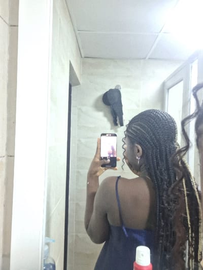 The image captures an indoor scene, located in the city of Close, Nigeria. A person is depicted from behind, reflected in a mirror, actively taking a selfie with a black smartphone held in their right hand. The individual has dark skin and long, dark braided hair, with some lighter strands visible within the braids. They are wearing a dark blue camisole with thin straps. A silver-colored earring is visible on their left ear.
The visible environment in the mirror's reflection features light-colored, patterned walls and a white acoustic panel ceiling. To the upper right of the person's reflection, a dark, human-figure-shaped object is mounted on the wall, appearing as a decorative hook or sculpture. On a surface in the lower foreground, a light blue pump bottle with a dispenser and a red and white aerosol can with a white nozzle are present. A vertical structural element or doorway is visible on the left side of the reflection, and a lighter, possibly windowed area is on the far right. Bright overhead lighting is evident, causing some glare on the mirror surface.