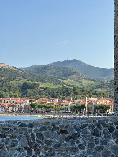 The image depicts a picturesque coastal village nestled against a backdrop of lush green mountains. A stone wall, likely part of a fortification or historic building, dominates the right side of the frame, its rough texture and irregular stones adding a sense of age and permanence. Through a gap in this wall, the scene unfolds.

In the mid-ground, a densely packed village with red-tiled roofs stretches along the coastline. The architecture suggests a Mediterranean setting, with buildings clustered closely together, some with balconies and varying shades of orange and peach facades. Trees, including palm trees, are interspersed among the buildings, indicating a warm climate. A clear blue sea laps at the shore, and a beach, populated by numerous small figures of people, can be seen. They appear to be enjoying a sunny day, with some likely swimming or sunbathing.

The mountains behind the village are covered in dense vegetation, with terraced sections suggesting cultivation, possibly vineyards or olive groves. The slopes are steep and dramatic, emphasizing the village's sheltered position. The sky above is a clear, light blue, with a few wispy clouds. The overall impression is one of a vibrant, sun-drenched holiday destination. The time of day appears to be midday or early afternoon, given the brightness and clear sky. The location is hinted to be Collioure, France, a well-known coastal town on the Mediterranean. No visible text is present in the image.
