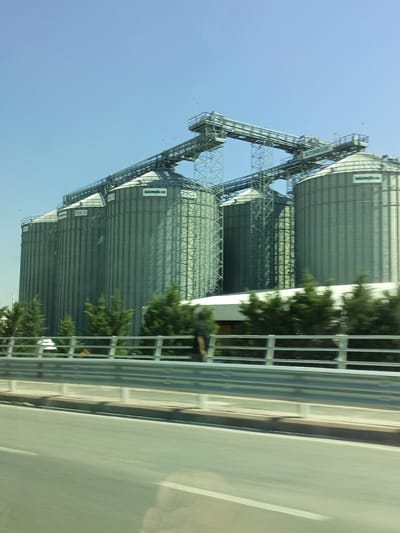 This media file captures an industrial scene with a row of large, silver grain silos under a clear, bright blue sky, indicating a sunny day. The silos are cylindrical and ribbed, with conical roofs. They are connected by a complex network of elevated walkways and chutes, suggesting an active transfer or storage facility. Green trees are visible in the foreground, partially obscuring the base of the silos and a metal guardrail that separates the industrial area from a road. The road itself is blurred due to motion, implying the photo was taken from a moving vehicle. The overall impression is one of industry and infrastructure, possibly a processing plant or storage depot. The lighting suggests it's daytime, likely mid-morning or afternoon. Some text is visible on the silos, appearing as white lettering against a dark green background. The visible text reads "BERKOGLU" and "BERKOGLU A.Ş." which suggests this is a facility belonging to Berkoglu A.Ş. While no specific event is depicted, the infrastructure points to ongoing operations related to grain storage or processing.